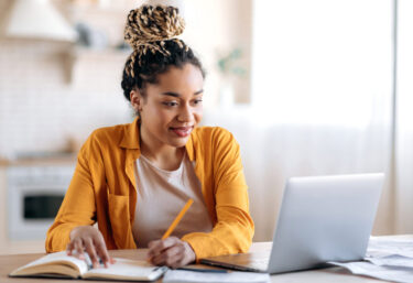 focused-cute-stylish-african-american-female-student-with-afro-dreadlocks-studying-remotely-from-home-using-a-laptop-taking-notes-on-notepad-during-online-lesson-e-learning-concept-smiling
