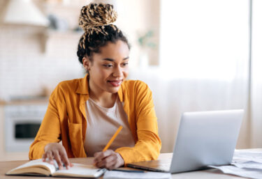 focused-cute-stylish-african-american-female-student-with-afro-dreadlocks-studying-remotely-from-home-using-a-laptop-taking-notes-on-notepad-during-online-lesson-e-learning-concept-smiling